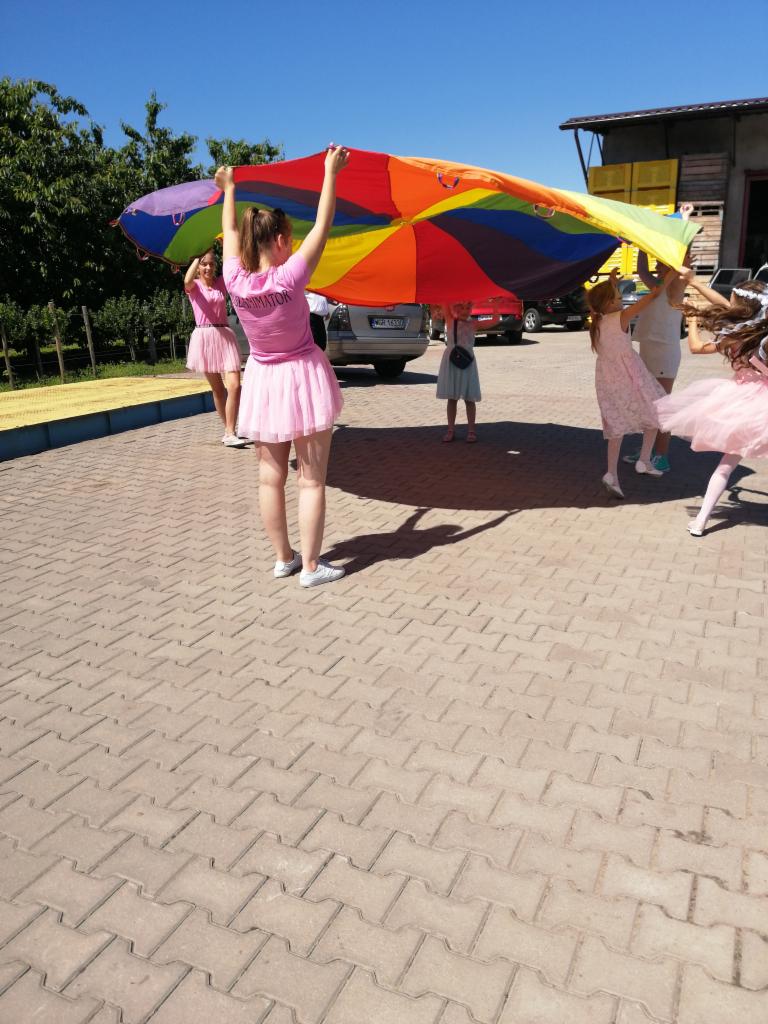 Animatorzy w różowych spódniczkach trzymają kolorową chustę animacyjną z dziećmi na zewnątrz, na tle budynków i samochodów.