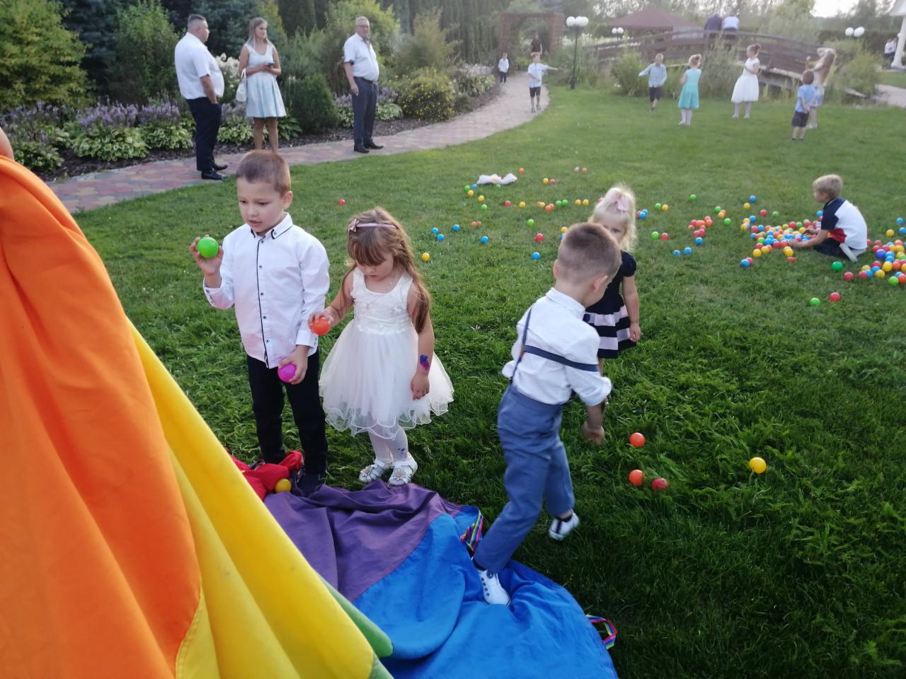 Dzieci bawiące się na trawie kolorowymi piłkami i pod kolorową chustą animacyjną podczas imprezy plenerowej, w tle widoczni dorośli.