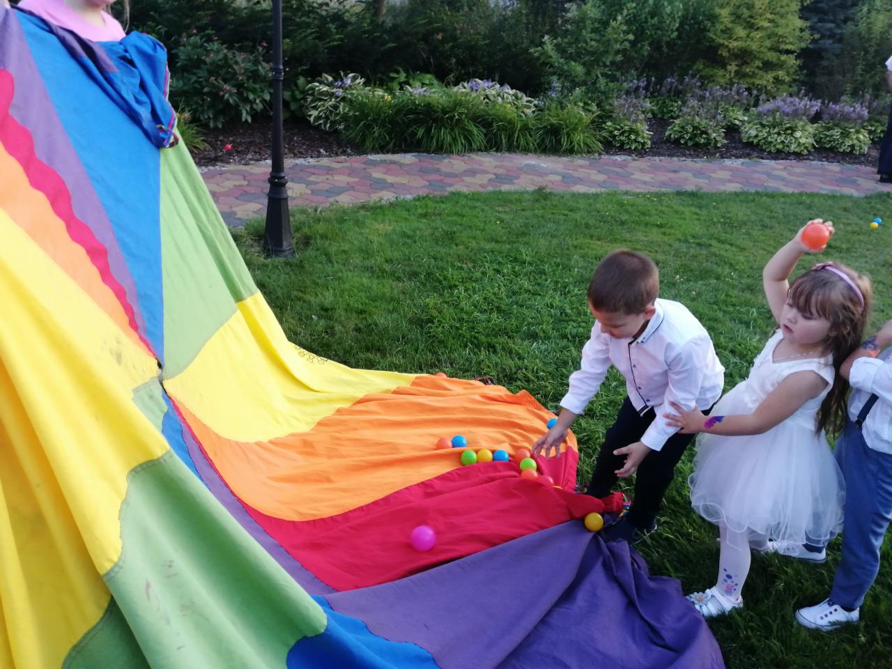 Dzieci w eleganckich strojach bawią się kolorową chustą animacyjną i piłeczkami na trawie podczas imprezy plenerowej.