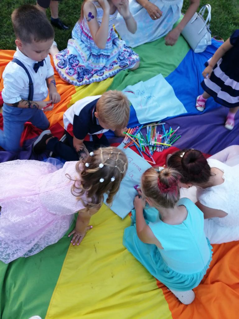 Grupa dzieci rysuje kredkami na kolorowej chuście podczas animacji, widoczny chłopiec z tatuażem na ręce i dziewczynki w sukienkach.