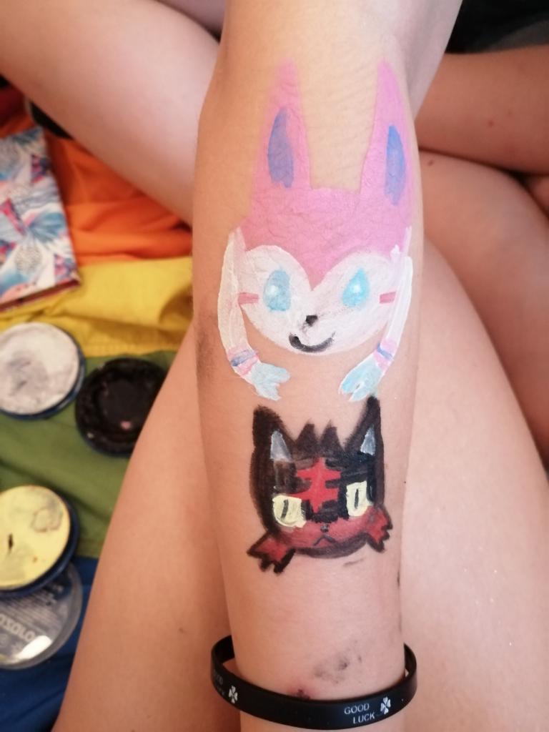 Ręka z pomalowanymi farbami postaciami z bajek, widoczne farby do malowania twarzy i tęczowe tło.