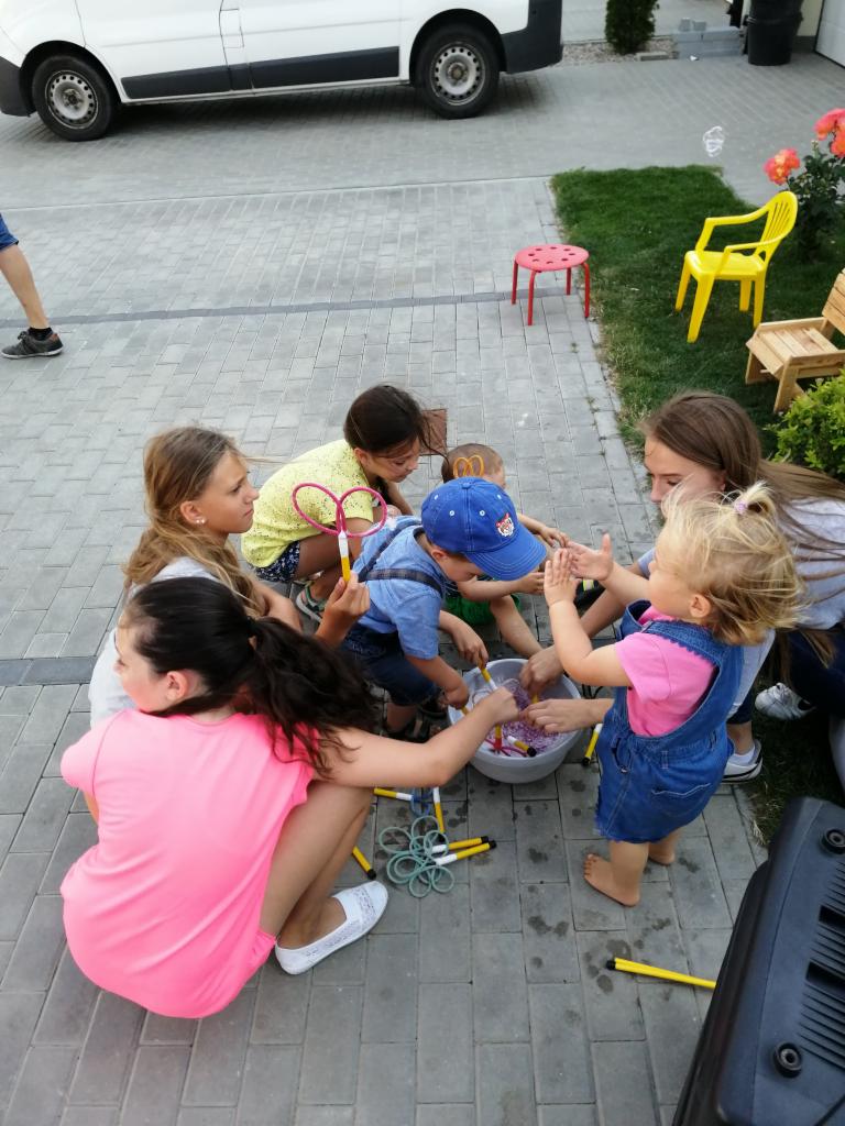 Grupa dzieci siedzi wokół miski z płynem do baniek mydlanych, używając żółtych narzędzi do robienia baniek, na zewnątrz, w tle zaparkowany biały samochód dostawczy.
