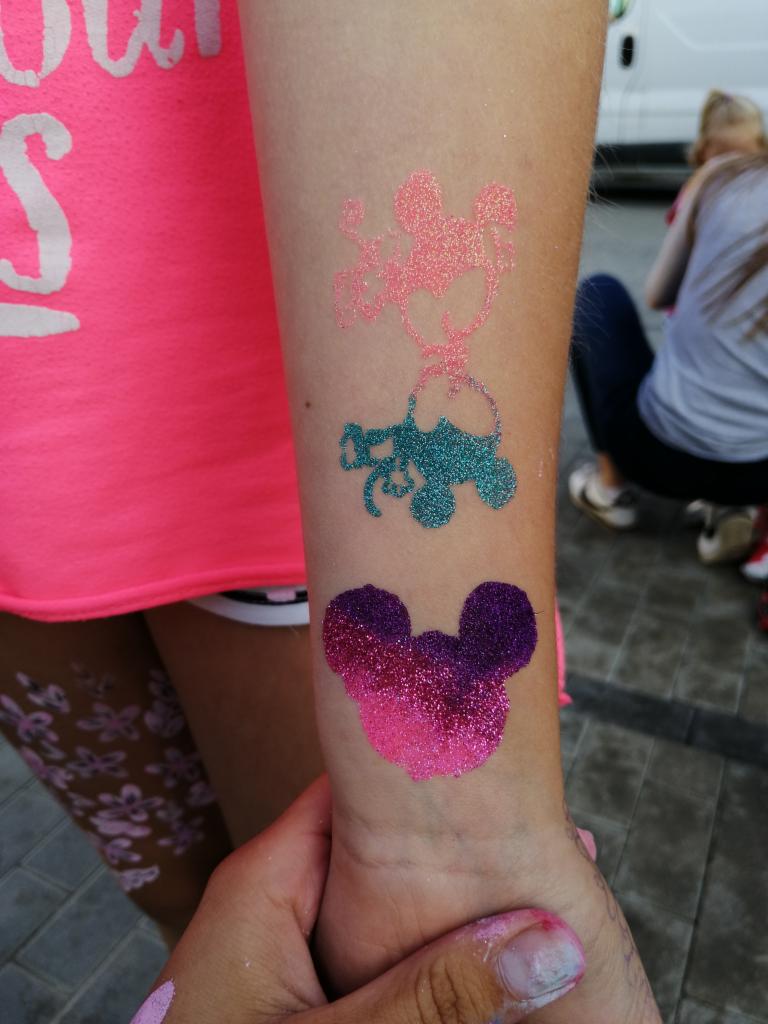 Ramię dziecka z brokatowymi tatuażami tymczasowymi: różowa Myszka Miki, turkusowy wózek i fioletowo-różowa Myszka Miki na nadgarstku.