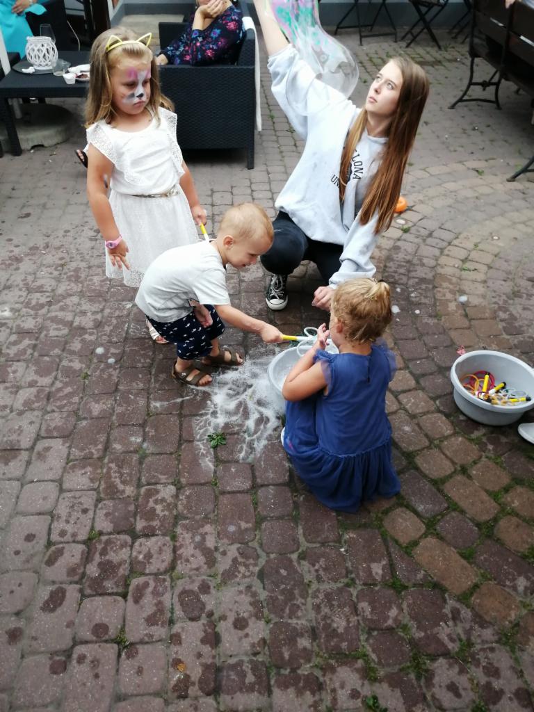 Dzieci bawią się w puszczanie baniek mydlanych na zewnątrz, dziewczynka z pomalowaną twarzą kota, starsza dziewczyna nadzoruje zabawę.