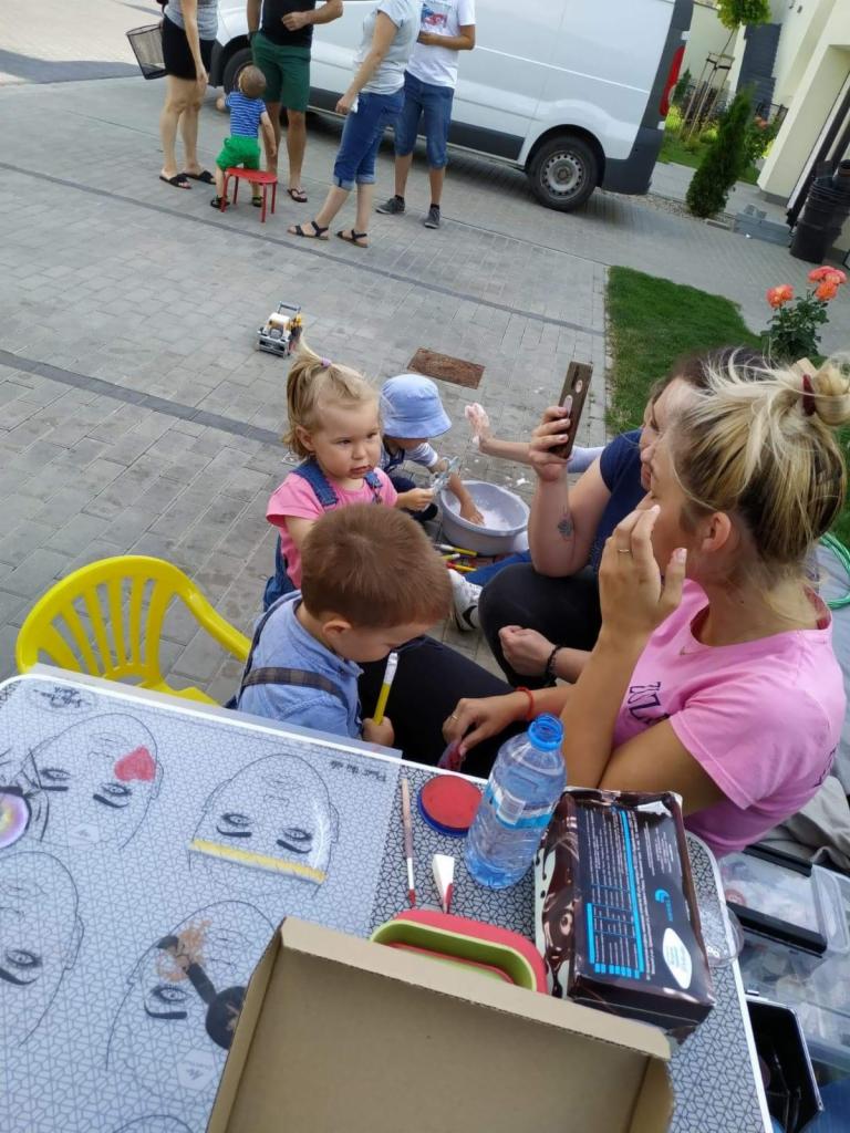 Dzieci podczas malowania twarzy na zewnątrz, widoczny stół z szablonami twarzy, pędzle, farby i butelka wody, w tle bawiące się dzieci i zaparkowany biały van.