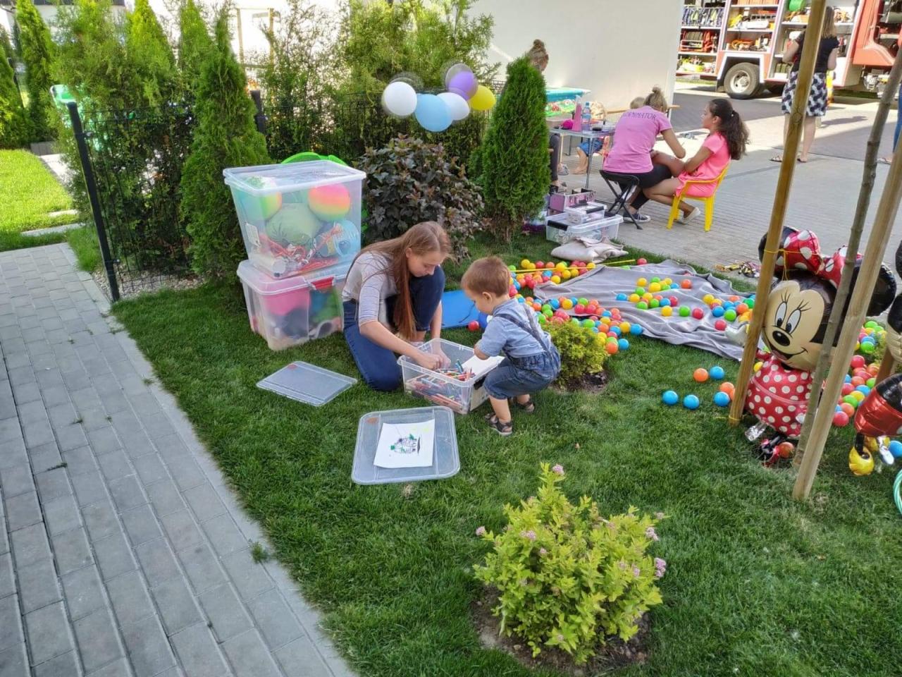 Animator dla dzieci podczas pracy: dziewczyna w dżinsach i mały chłopiec siedzą na trawie, obok nich pojemniki z zabawkami i kolorowe piłki, w tle wóz strażacki.