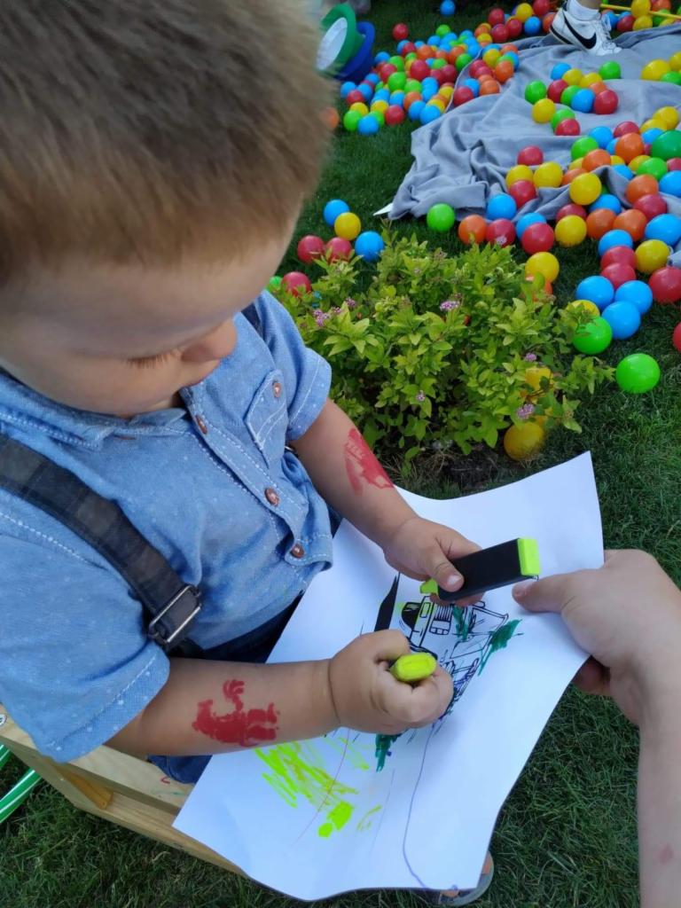 Chłopiec z tymczasowymi tatuażami na ramionach rysuje żółtym markerem na kartce, obok kolorowe piłeczki w tle.