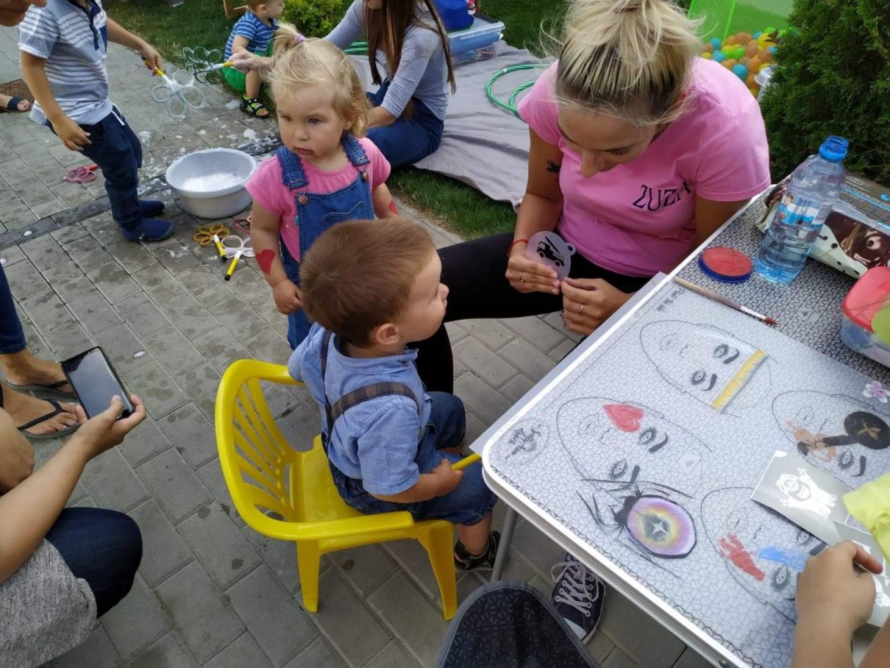 Animator maluje twarz dziecka podczas imprezy plenerowej, w tle dzieci puszczają bańki mydlane.