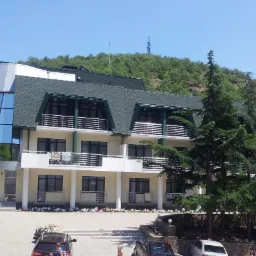 Nowoczesny budynek z elewacją w jasnych kolorach i dużymi oknami, otoczony zielenią, z widoczną g&oacute;rą w tle.