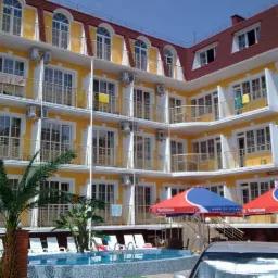 Żółty budynek hotelowy z balkonami, basenem, palmami i parasolami, widok z poziomu ulicy, częściowo zasłonięty samochodem.