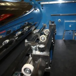 Fragment niebieskiej maszyny introligatorskiej z czarnymi rolkami i metalowymi elementami mocującymi.