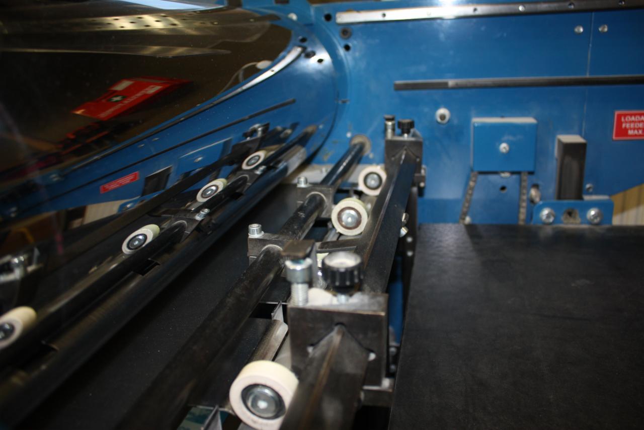 Fragment niebieskiej maszyny introligatorskiej z czarnymi rolkami i metalowymi elementami mocującymi.