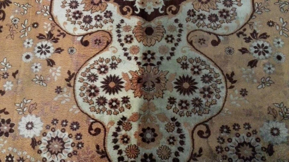 Wzór kwiatowy na beżowym dywanie, widoczne plamy i zabrudzenia na jasnym tle.