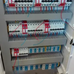 Rozdzielnica elektryczna z zainstalowanymi bezpiecznikami i przewodami w kolorach czerwonym i niebieskim, widoczne trzy rzędy zabezpieczeń.