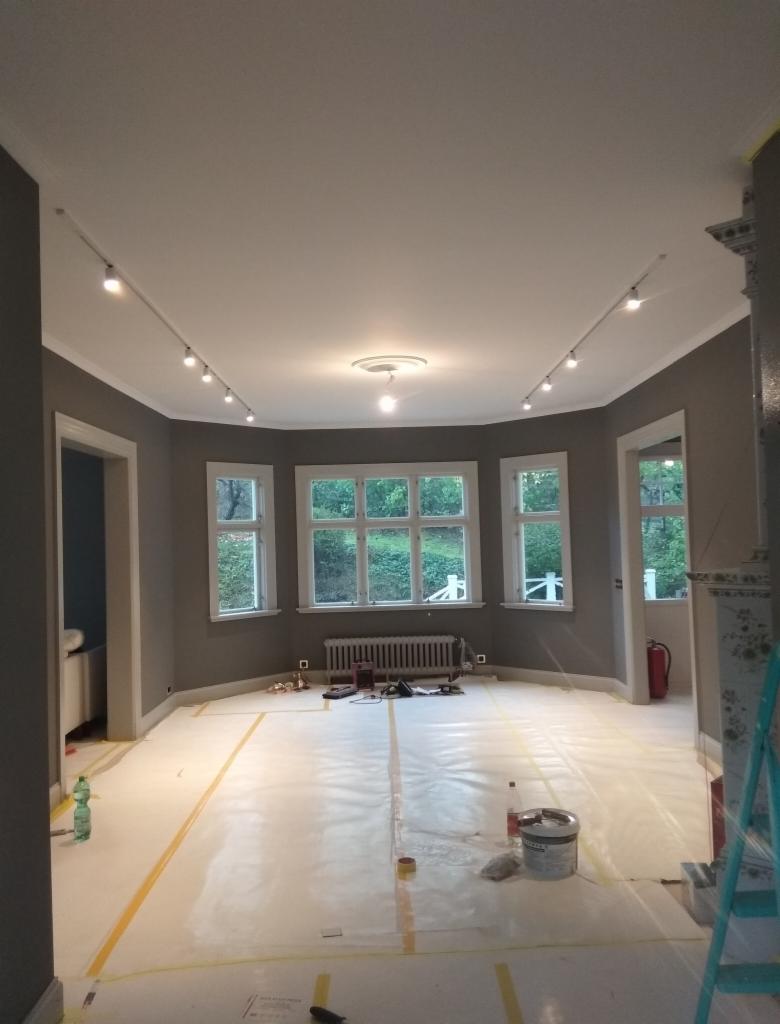 Pokój w trakcie przygotowań do malowania: podłoga zabezpieczona folią, ściany w szarym kolorze, widoczne puszki z farbą i narzędzia, listwa oświetleniowa na suficie.