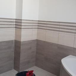 Andrzej Trzeciak - Wykończona łazienka z płytkami imitującymi drewno, pralką i częściowo widoczną białą wanną, widoczne wystające rury ciepłej i zimnej wody.