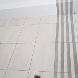 Andrzej Trzeciak - Ściana wyłożona jasnymi, drewnopodobnymi płytkami z dekoracyjnym pionowym pasem szarych płytek, widoczny wystający przewód elektryczny i fragment odpływu podłogowego.