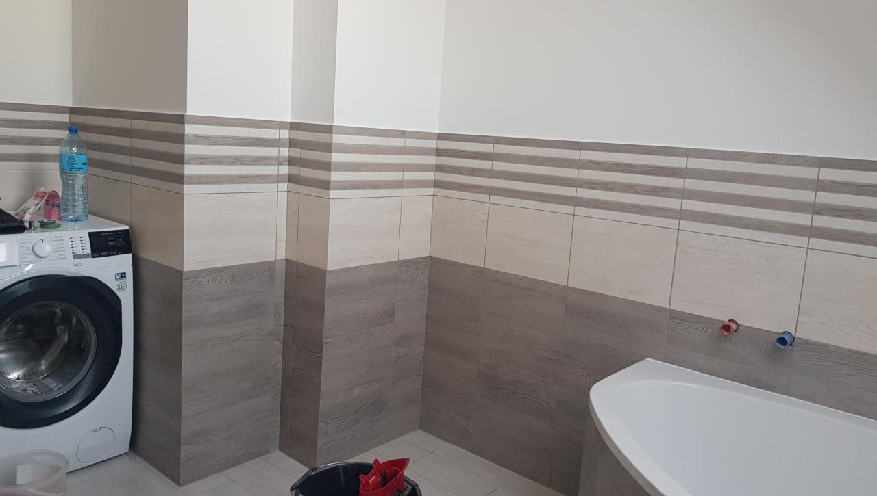 Wykończona łazienka z płytkami imitującymi drewno, pralką i częściowo widoczną białą wanną, widoczne wystające rury ciepłej i zimnej wody.