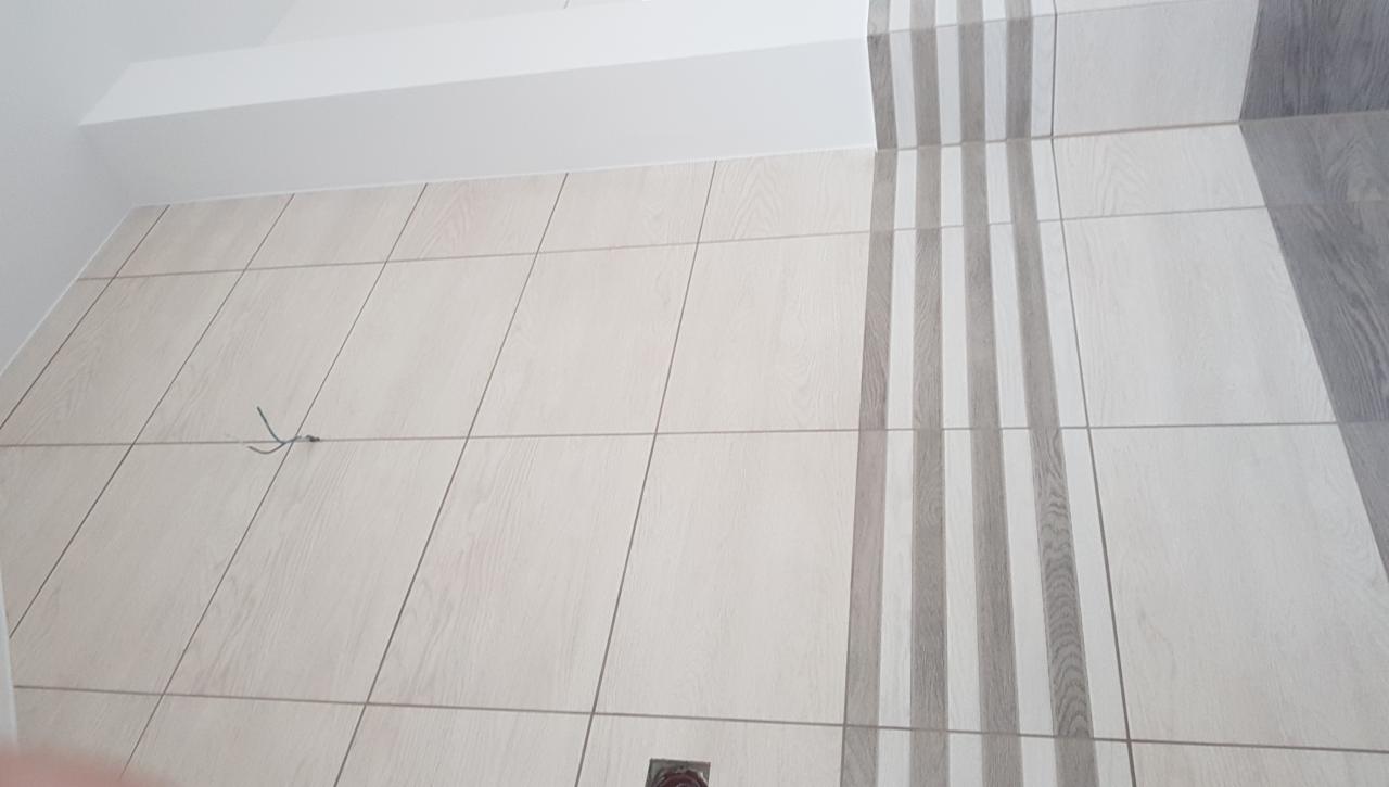 Ściana wyłożona jasnymi, drewnopodobnymi płytkami z dekoracyjnym pionowym pasem szarych płytek, widoczny wystający przewód elektryczny i fragment odpływu podłogowego.