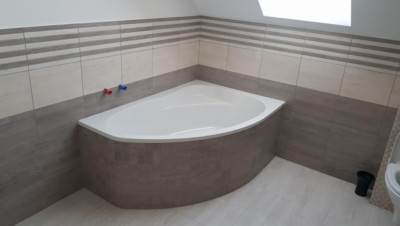 Narożna wanna obudowana płytkami w odcieniach szarości i beżu, z widocznymi wystającymi rurami z ciepłą i zimną wodą oraz fragmentem toalety w tle.