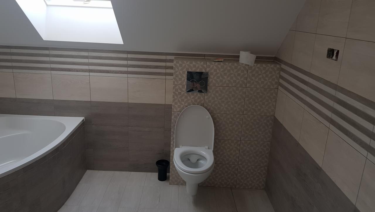 Ukończona łazienka na poddaszu ze skośnym sufitem, z wanną, toaletą i wykończoną glazurą w odcieniach beżu i szarości, widoczne niedociągnięcia w postaci braku listwy przy podłodze.