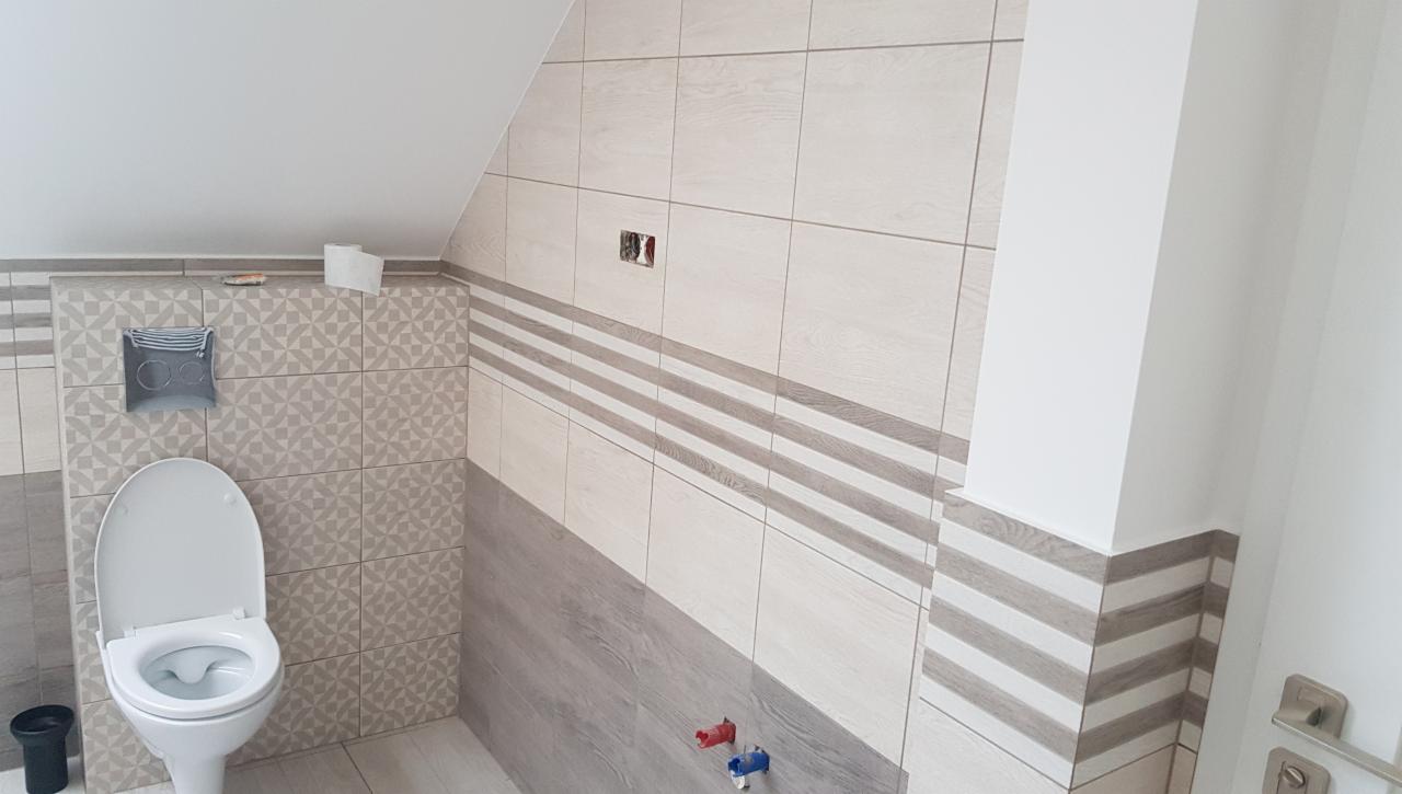 Wykończona łazienka na poddaszu z toaletą, wzorzystymi płytkami i poziomymi pasami dekoracyjnymi, widoczne przyłącza wodne i otwór na gniazdko elektryczne.