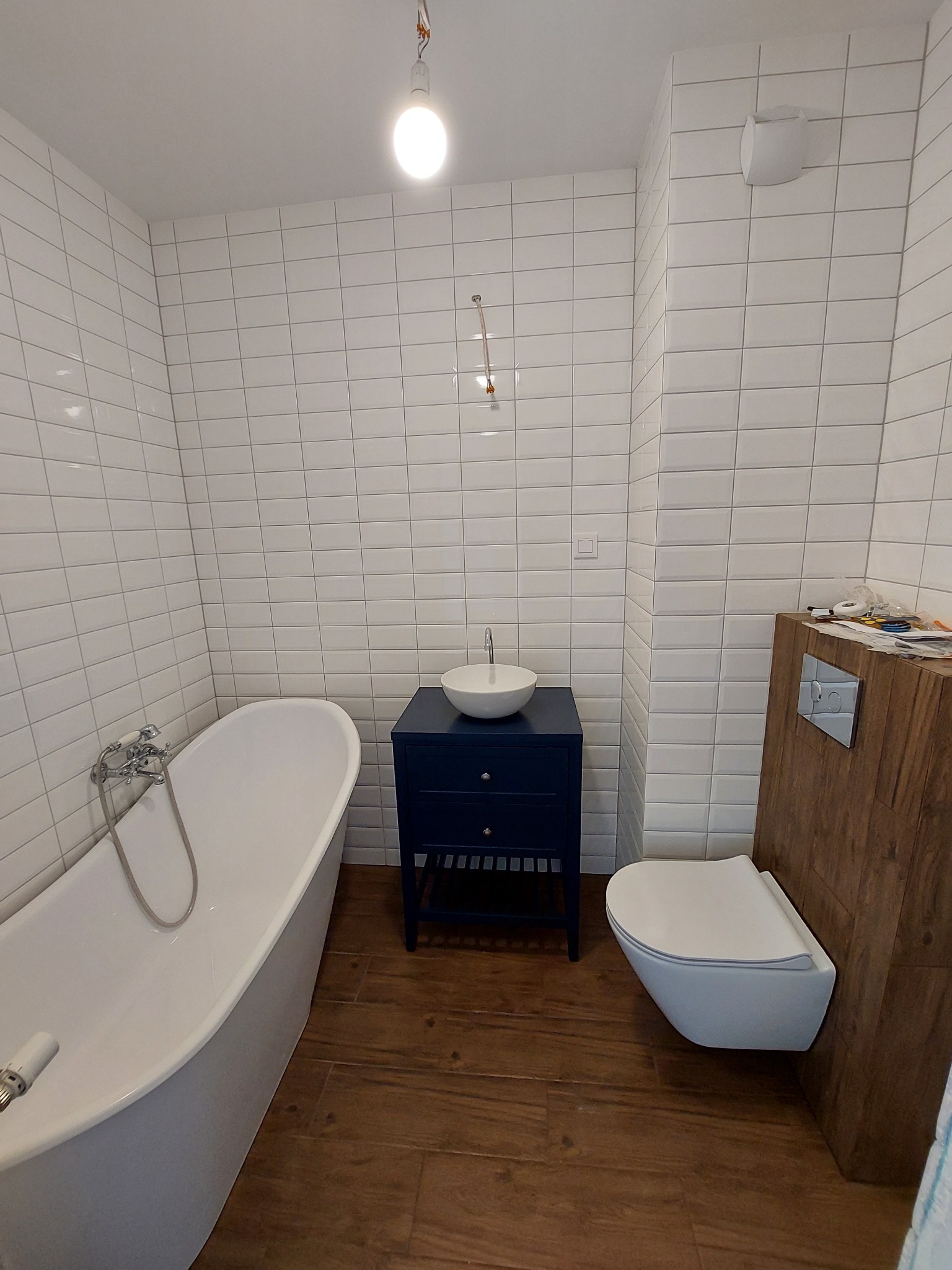 Białe, prostokątne płytki ścienne w łazience z wanną, umywalką nablatową na granatowej szafce i podwieszanym WC z drewnianą obudową stelaża.