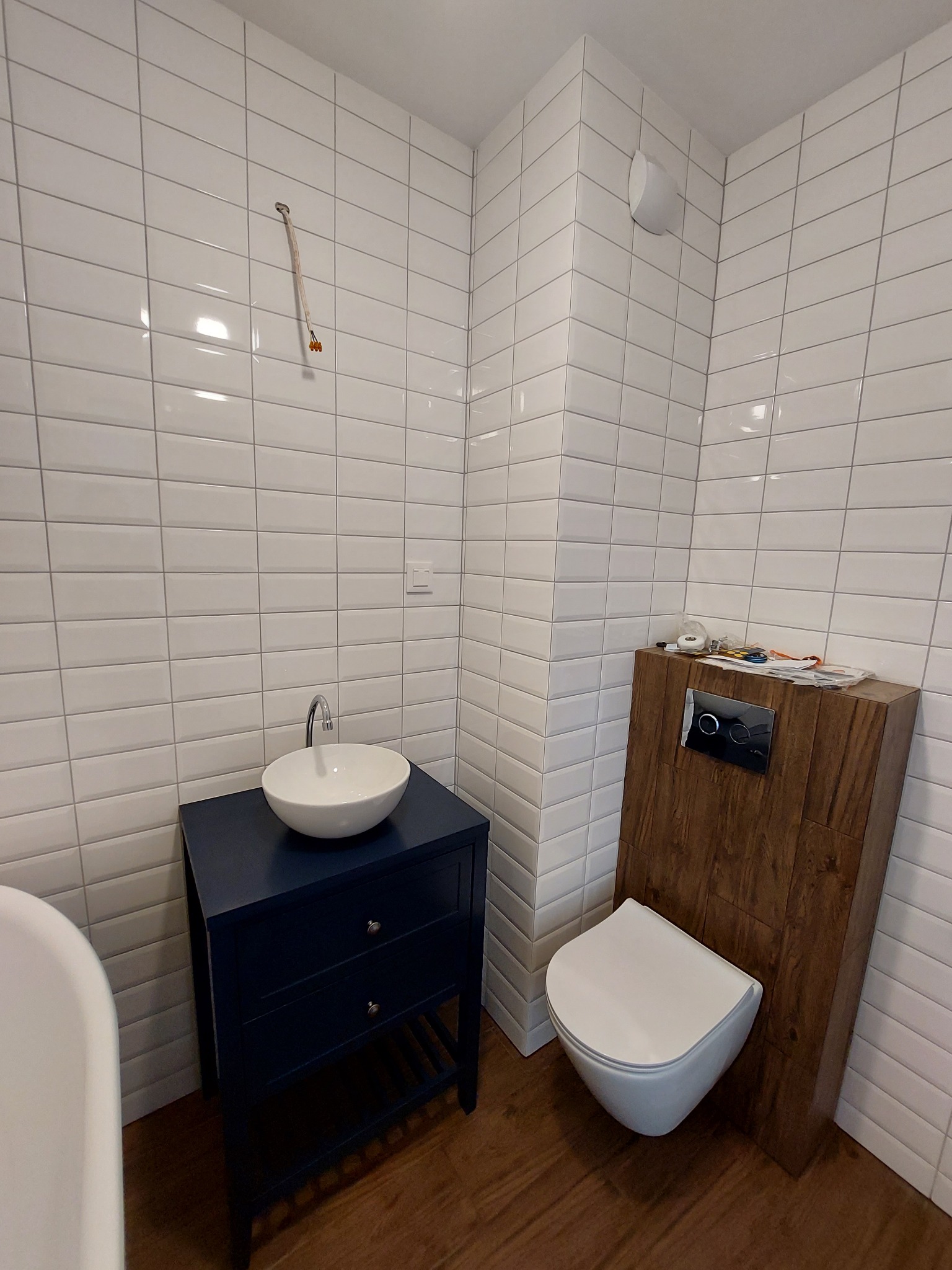 Nowoczesna łazienka z białymi, prostokątnymi płytkami, ciemnoniebieską szafką pod umywalką z okrągłą, białą umywalką nablatową, wiszącą toaletą z obudową z paneli drewnopodobnych i akcesoriami...