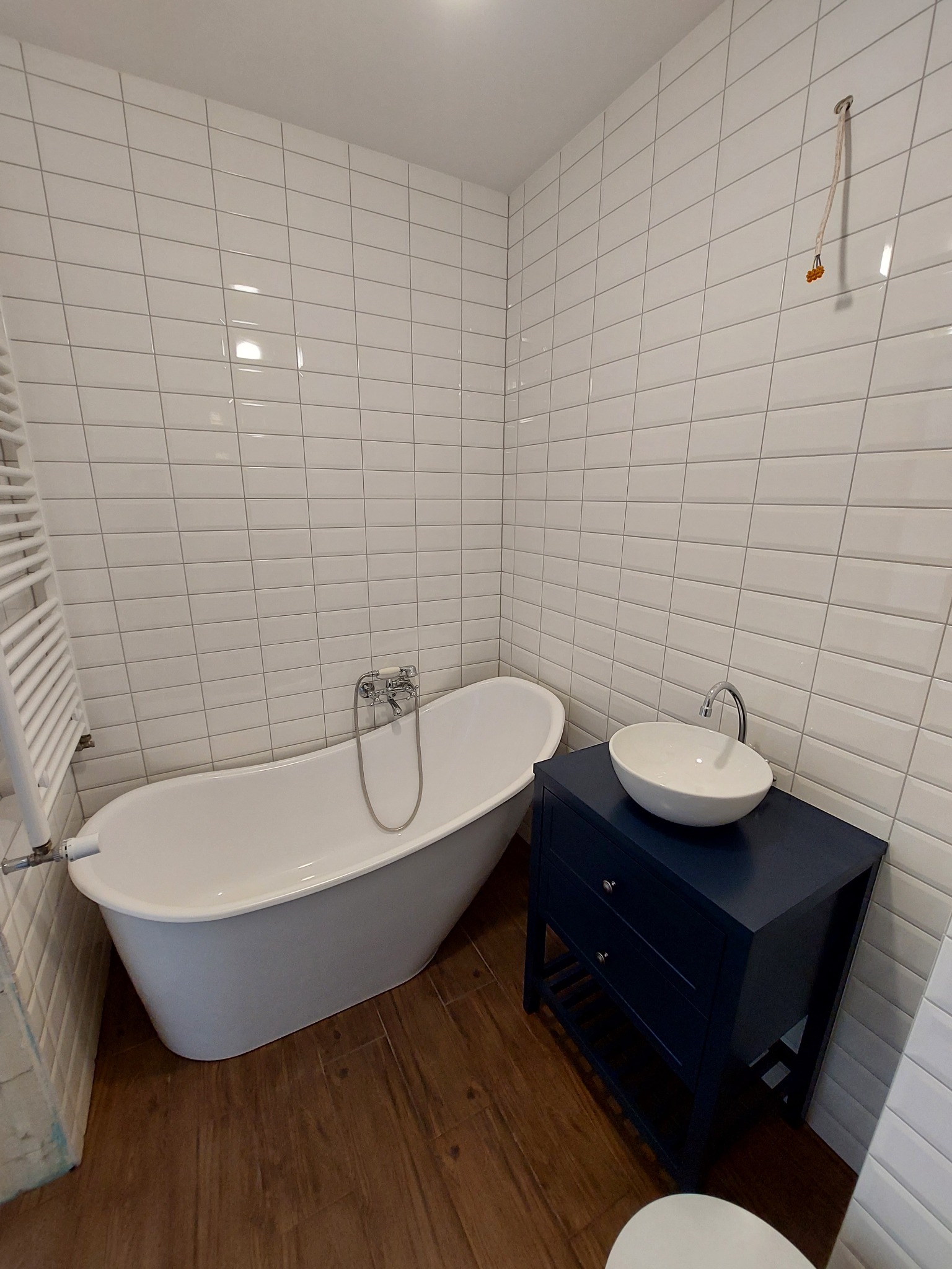 Biała wanna wolnostojąca z chromowaną armaturą obok umywalki nablatowej na granatowej szafce w łazience wyłożonej białymi, prostokątnymi płytkami.