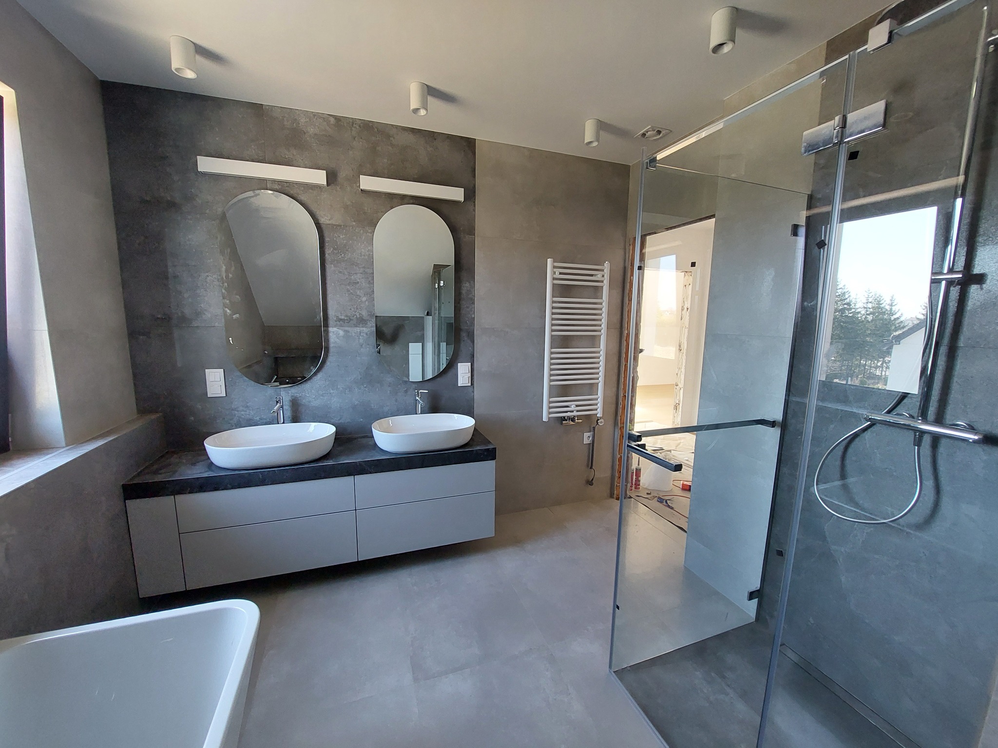 Nowoczesna łazienka z szarymi płytkami, owalnymi umywalkami nablatowymi i kabiną prysznicową typu walk-in, widok na częściowo wykończone wnętrze przez szklaną ścianę.