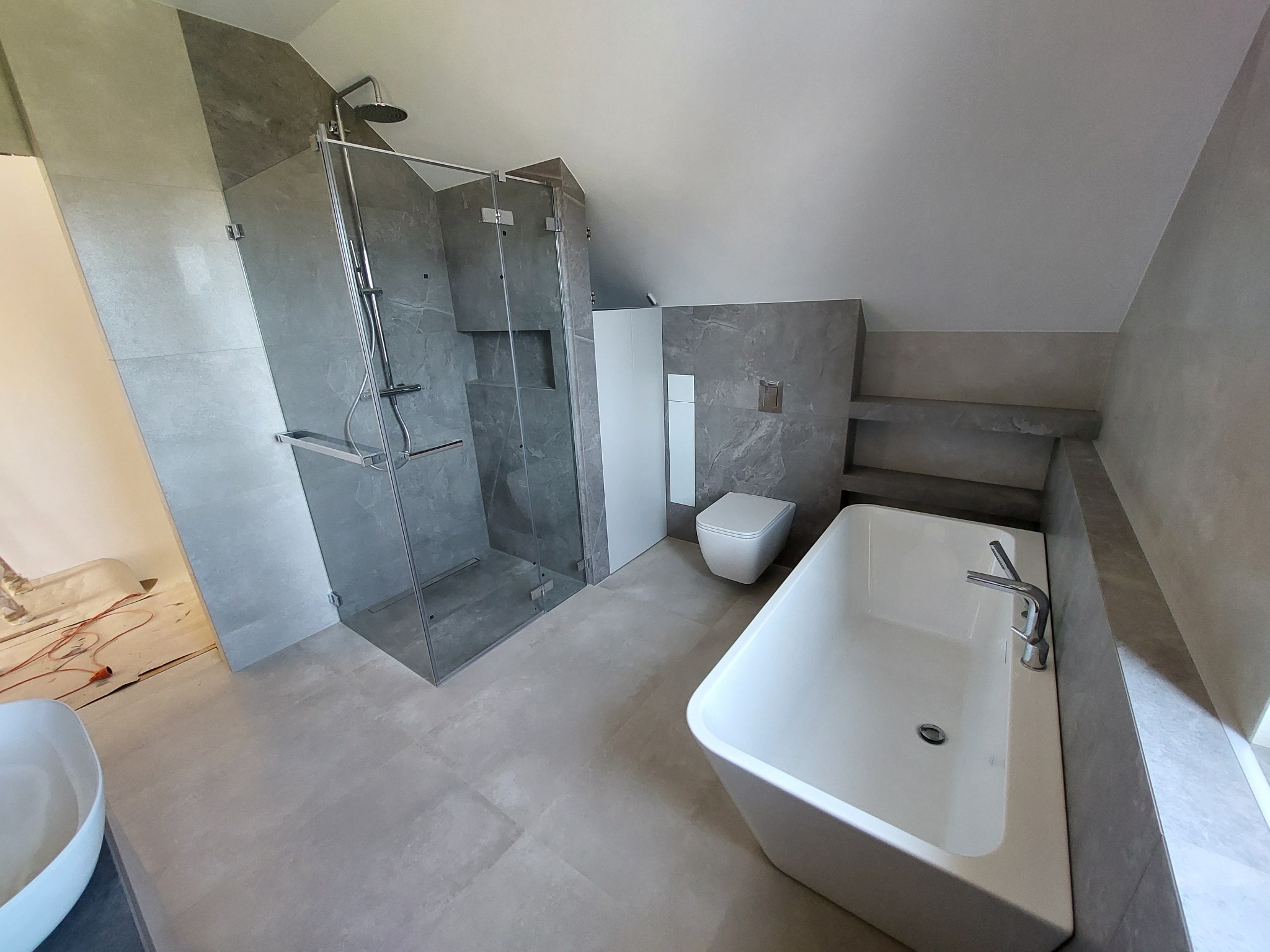 Wykończona łazienka na poddaszu z szarą, marmurkową glazurą, kabiną prysznicową typu walk-in i białą wanną prostokątną, oświetloną naturalnym światłem.