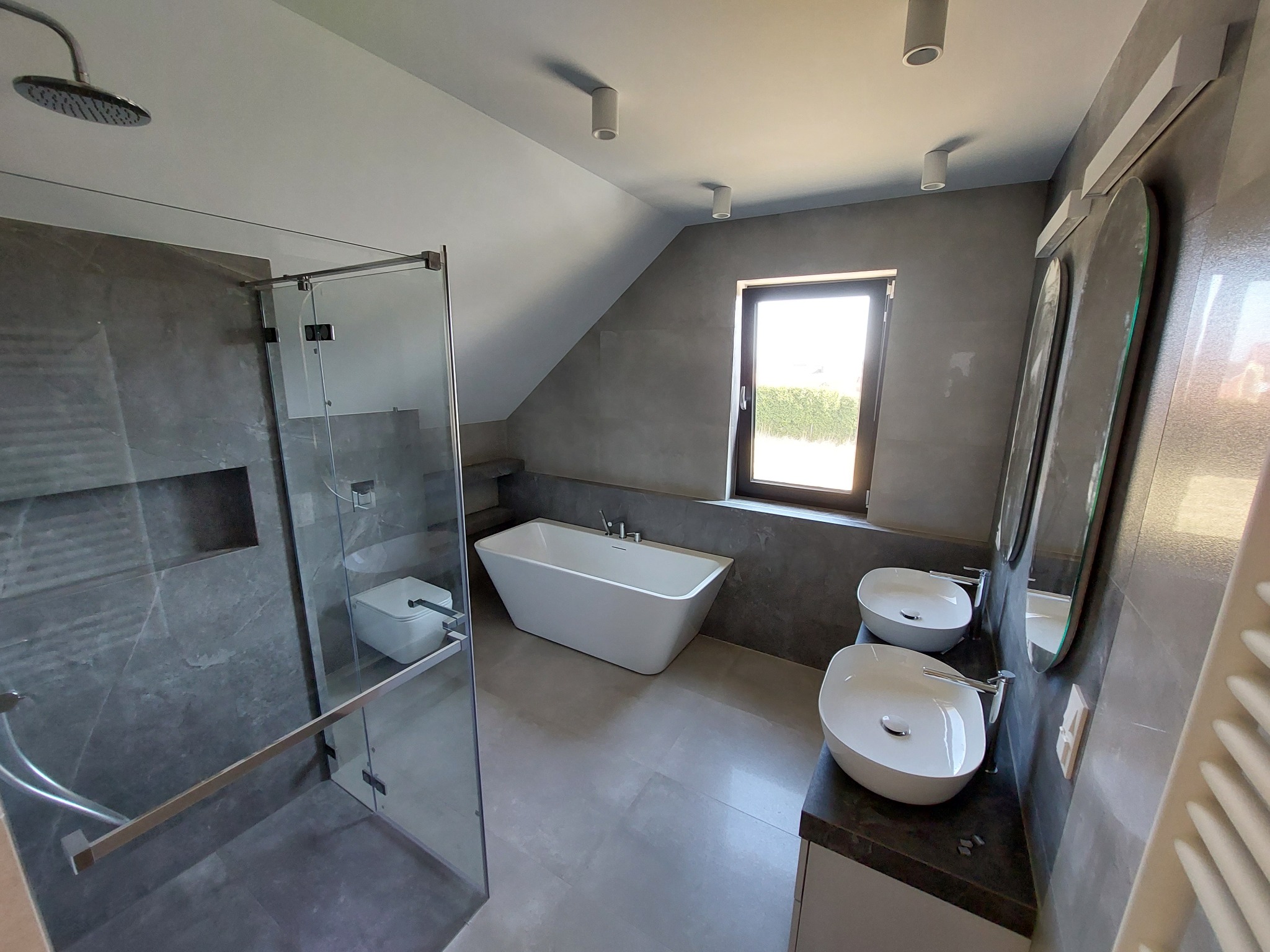 Nowoczesna łazienka w szarościach z kabiną prysznicową ze szklanymi drzwiami, wolnostojącą białą wanną, dwiema umywalkami nablatowymi i dużym oknem.