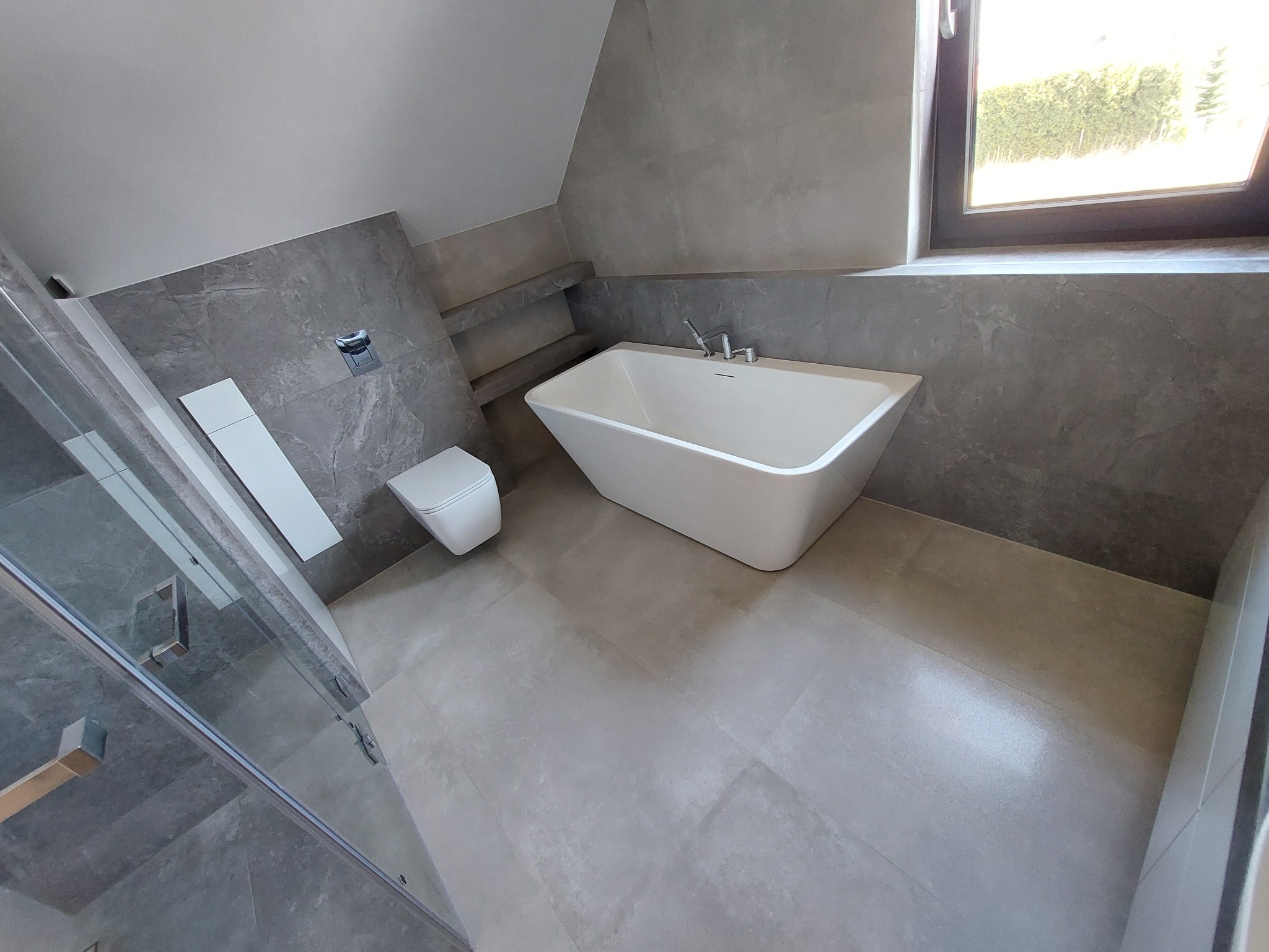 Nowoczesna łazienka w odcieniach szarości z wolnostojącą wanną, podwieszaną toaletą i kabiną prysznicową ze szklanymi drzwiami, wykończona płytkami imitującymi kamień, oświetlona naturalnym...
