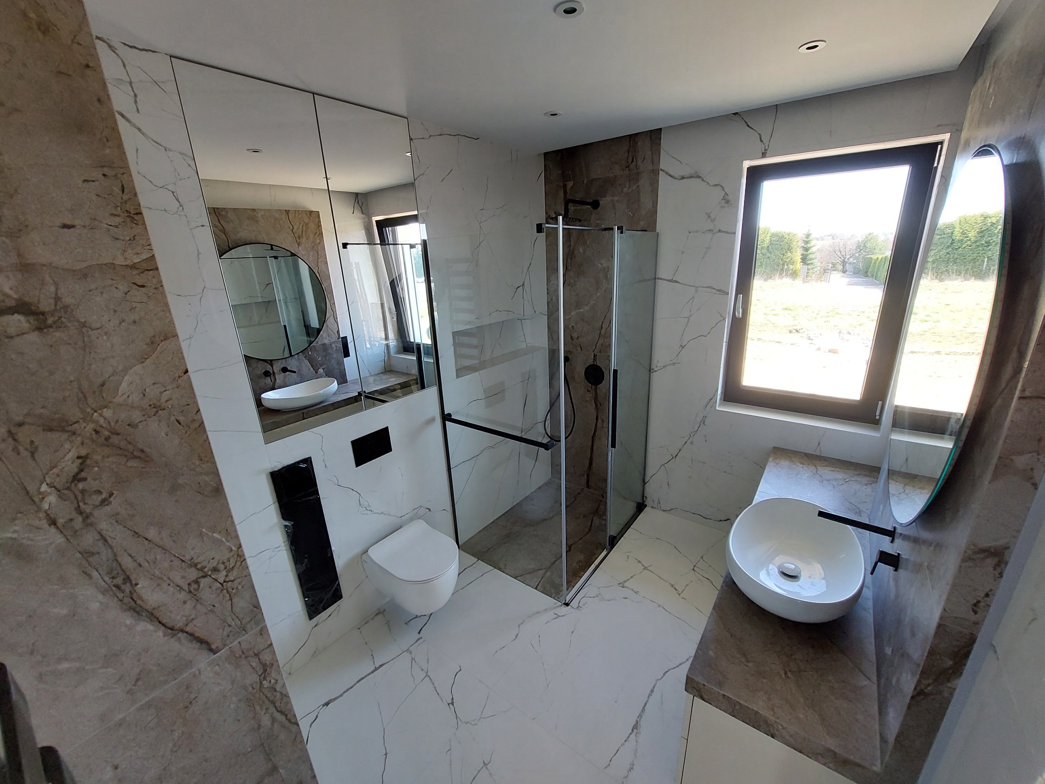 Nowoczesna łazienka z marmurowymi płytkami, kabiną prysznicową typu walk-in z czarnymi okuciami, wiszącą toaletą i umywalką nablatową o owalnym kształcie