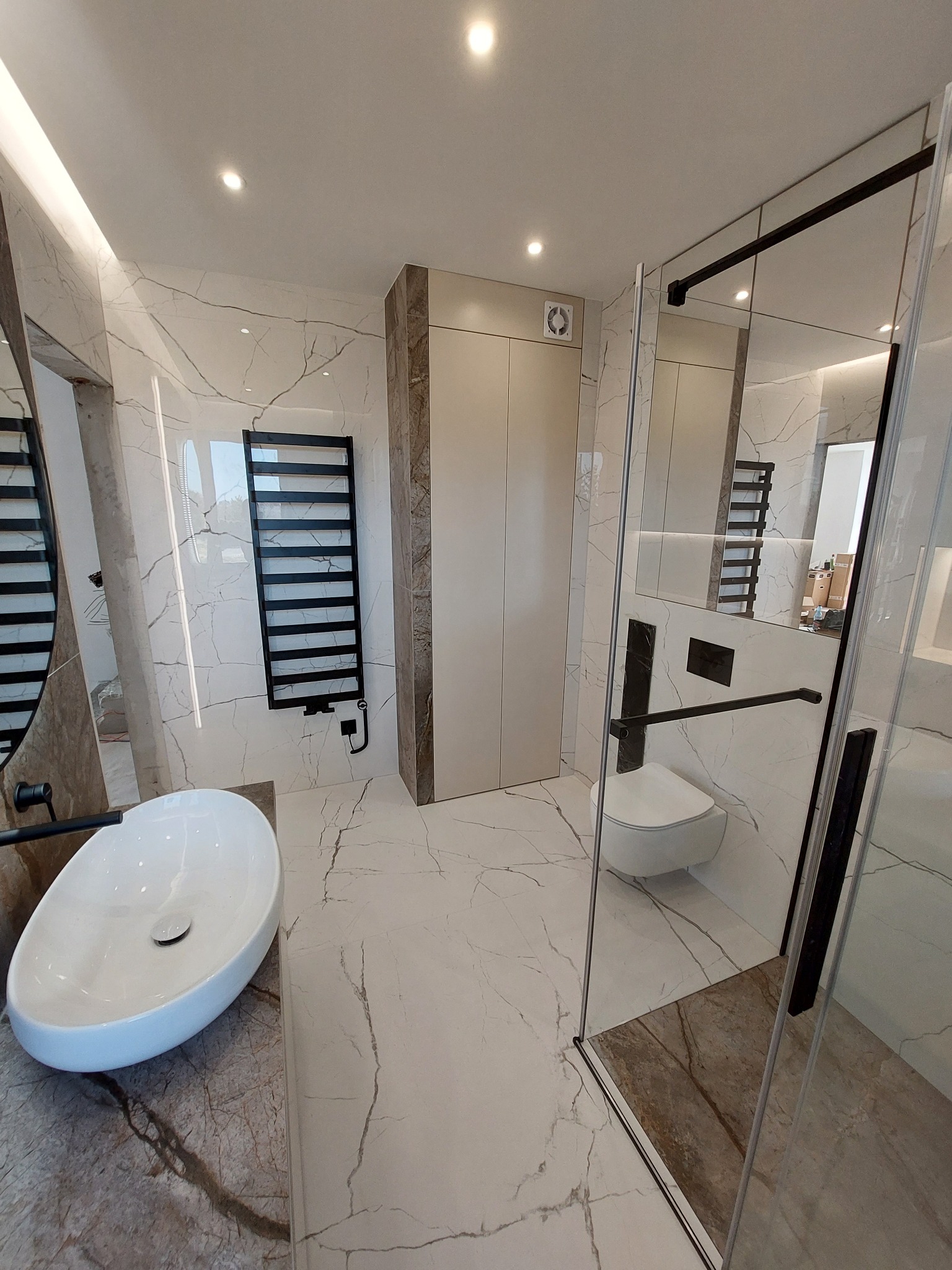 Nowoczesna łazienka z białymi marmurowymi płytkami, owalną umywalką nablatową na blacie z brązowego marmuru, czarnym grzejnikiem drabinkowym i kabiną prysznicową z czarnymi profilami.