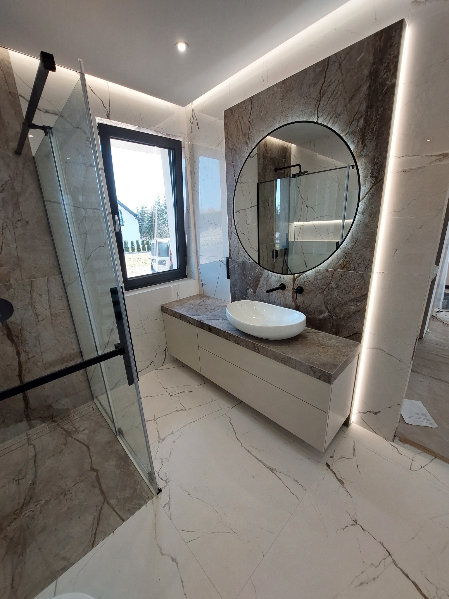 Nowoczesna łazienka z marmurowymi płytkami, okrągłym lustrem z podświetleniem LED, białą umywalką nablatową i kabiną prysznicową ze szklanymi drzwiami z czarnymi ramami.