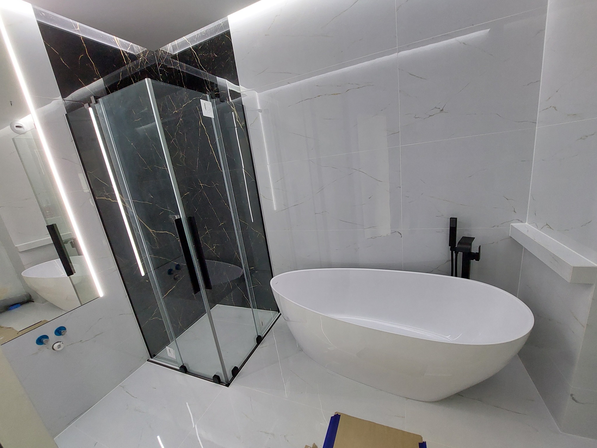 Nowoczesna łazienka z czarno-białymi marmurkowymi płytkami, kabiną prysznicową typu walk-in z czarnymi profilami i wolnostojącą białą wanną owalną, oświetlenie LED w suficie.