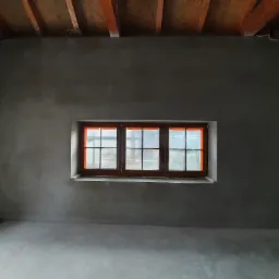 Surowe wnętrze z oknem zabezpieczonym folią, ściany pokryte świeżym tynkiem, widoczne drewniane elementy konstrukcji dachu.