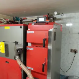 Dwa kotły grzewcze, jeden szary z żółtymi naklejkami, drugi czerwony z czarnymi uchwytami, połączone rurą, zainstalowane w pomieszczeniu z kafelkami na ścianach.