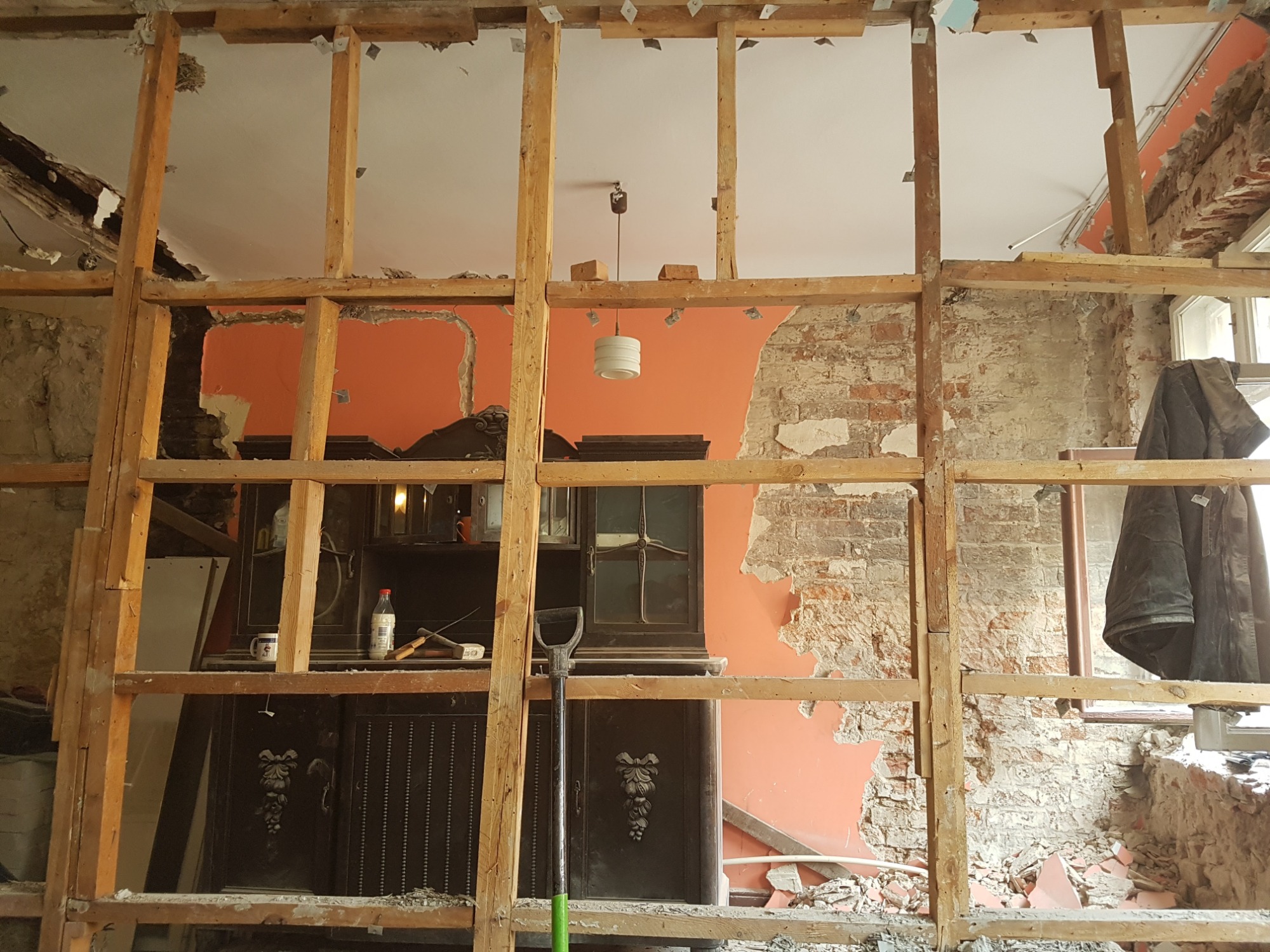 Demontaż ściany działowej z widoczną starą cegłą, fragmentami pomarańczowej ściany i starą szafą w tle, drewniana konstrukcja stelażu w pierwszym planie.