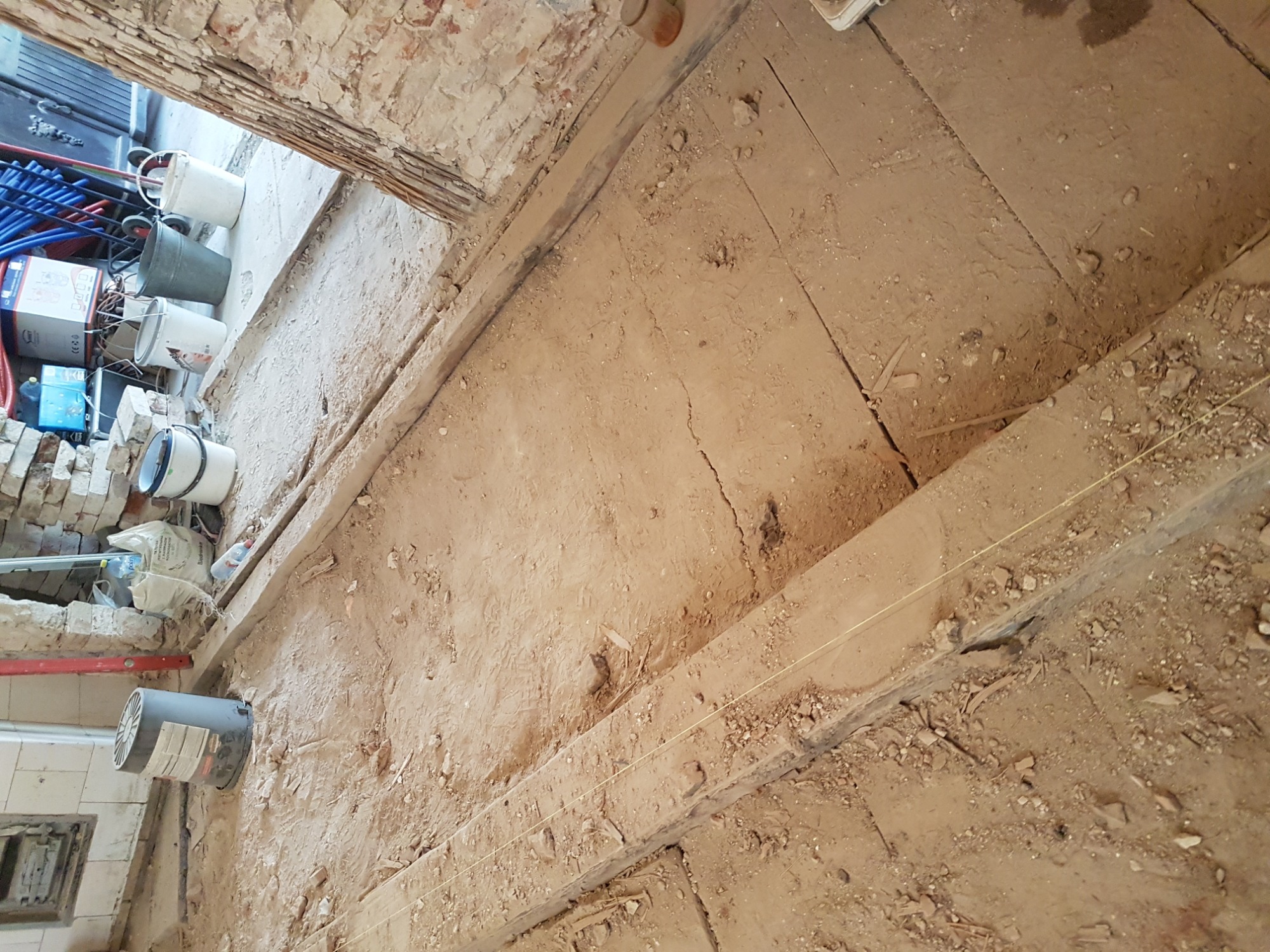 Podłoga w trakcie remontu w Toruniu, widoczne stare deski, gruz, ściany z cegły i przygotowane wiadra z materiałami budowlanymi.