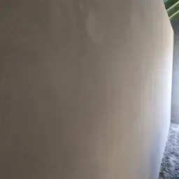 Świeżo nałożony, jasnoszary tynk maszynowy na ścianie wewnętrznej, widoczne zielone elementy konstrukcji dachu w tle, surowy stan wykończenia podłogi.