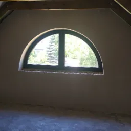 Poddasze po nałożeniu gładzi szpachlowej, widoczne drewniane krokwie i okno w kształcie półkola z widokiem na zieleń.