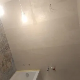 Ściany łazienki wyłożone jasnymi płytkami ceramicznymi, częściowo z dekoracyjnym wzorem, widoczne wystające kable i rury, przygotowane do montażu armatury.