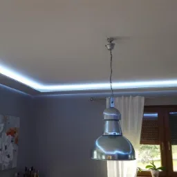 Pokój z oświetleniem LED podsufitowym, widoczna lampa wisząca w stylu industrialnym, obraz na ścianie i okno z zasłonami.