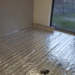 Montaż ogrzewania podłogowego: rury grzewcze ułożone na folii aluminiowej w przygotowanym pomieszczeniu, widoczne rękawice robocze.