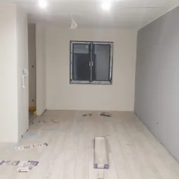 Pokój w trakcie remontu: świeżo położona jasna podłoga, pomalowana szara ściana, okno z czarną ramą, widoczne narzędzia i materiały budowlane