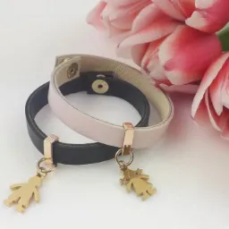 Dwa skórzane bransoletki: czarna z zawieszką chłopca i różowa z zawieszką dziewczynki, leżące na białym tle z rozmytymi różowymi tulipanami w tle.