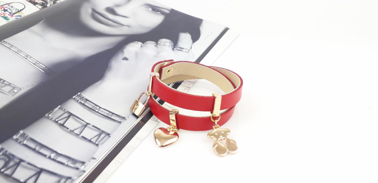 Czerwona, skórzana bransoletka z zawieszkami w kształcie serca, misia i kłódki, leżąca na okładce magazynu z czarno-białym portretem kobiety z biżuterią.