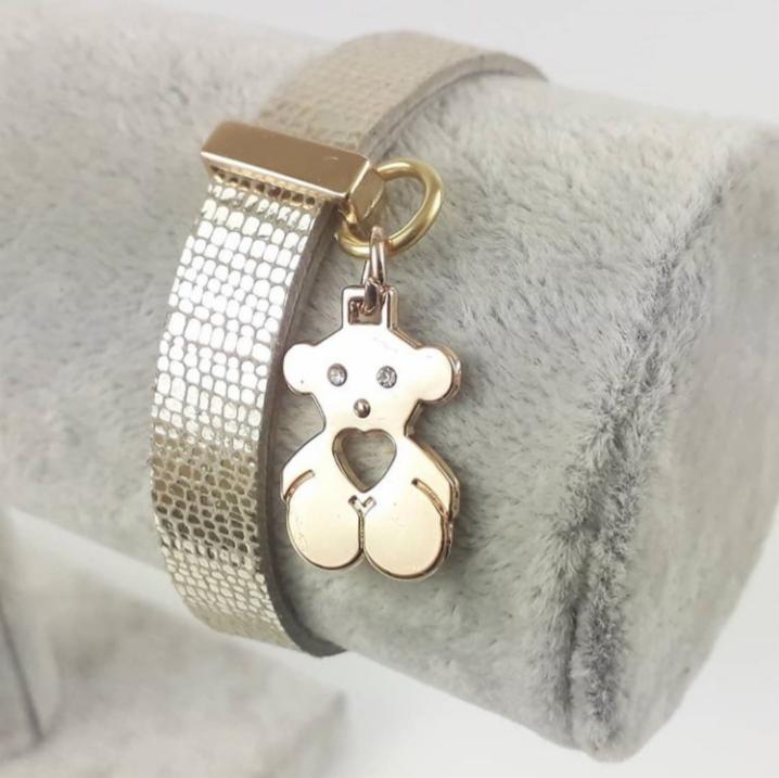 Bransoletka ze złotą zawieszką w kształcie misia z sercem, leżąca na szarym, pluszowym podłożu.